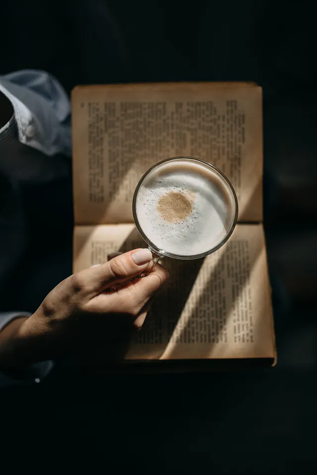 Une main tenant une tasse de boisson chaude au-dessus d'un livre ouvert, lumière douce